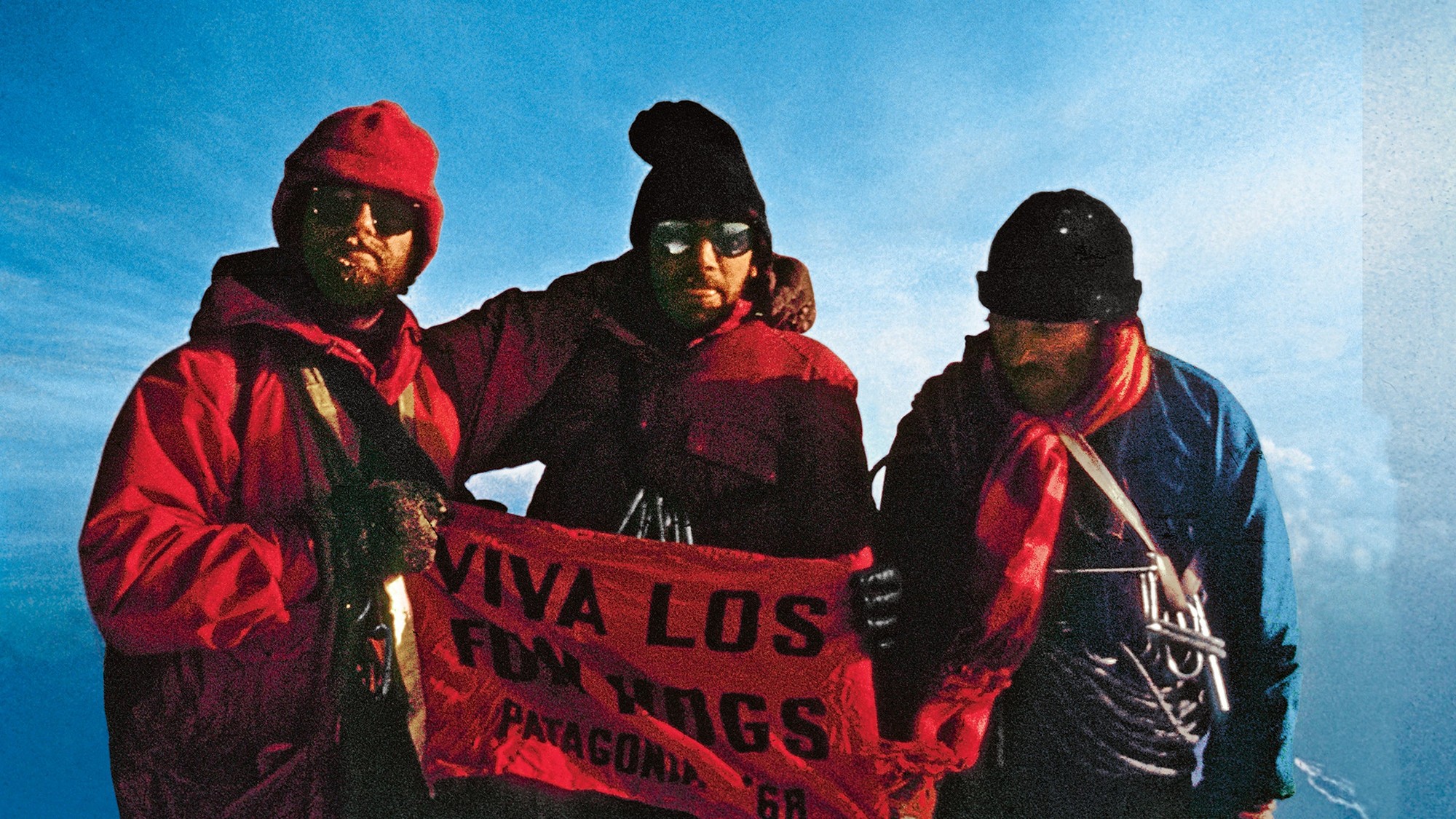 Três escaladores com roupa vermelha de frio, gorro e óculos escuros seguram uma faixa vermelha no topo de uma montanha, contra o céu azul