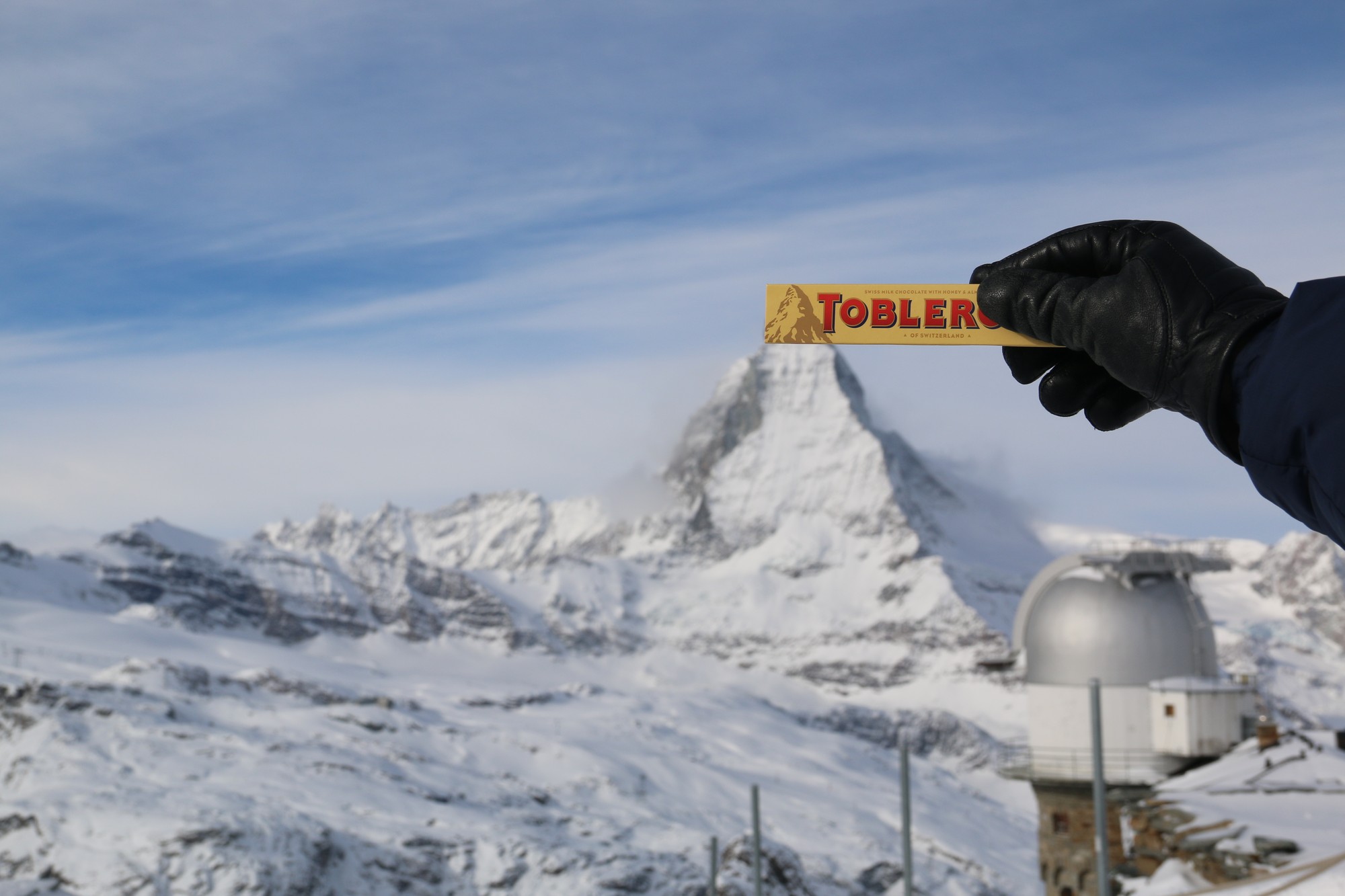 Uma mão de luva preta segura uma barra de Toblerone com a embalagem antiga no alto da foto, alinhada com o pico nevado do Matterhorn ao fundo
