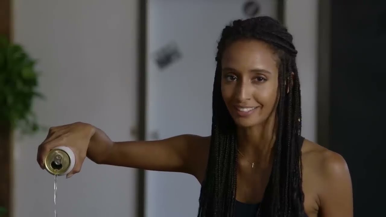 Uma mulher sorri pra câmera enquanto derrama água de uma lata, numa cena do comercial