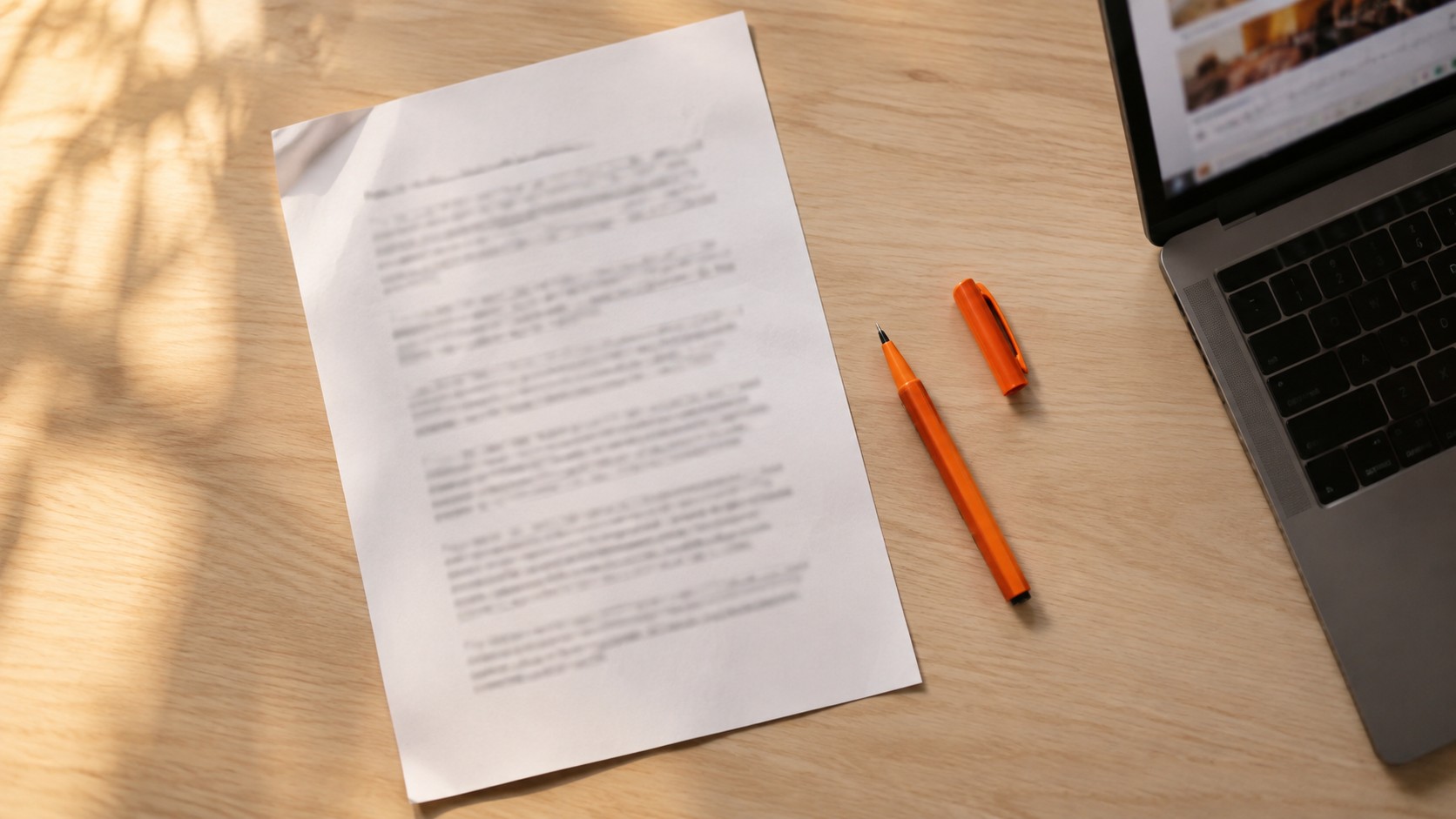 Folha impressa com parágrafos desfocados sobre uma mesa de trabalho, com uma caneta laranja do lado e um notebook aberto ao fundo