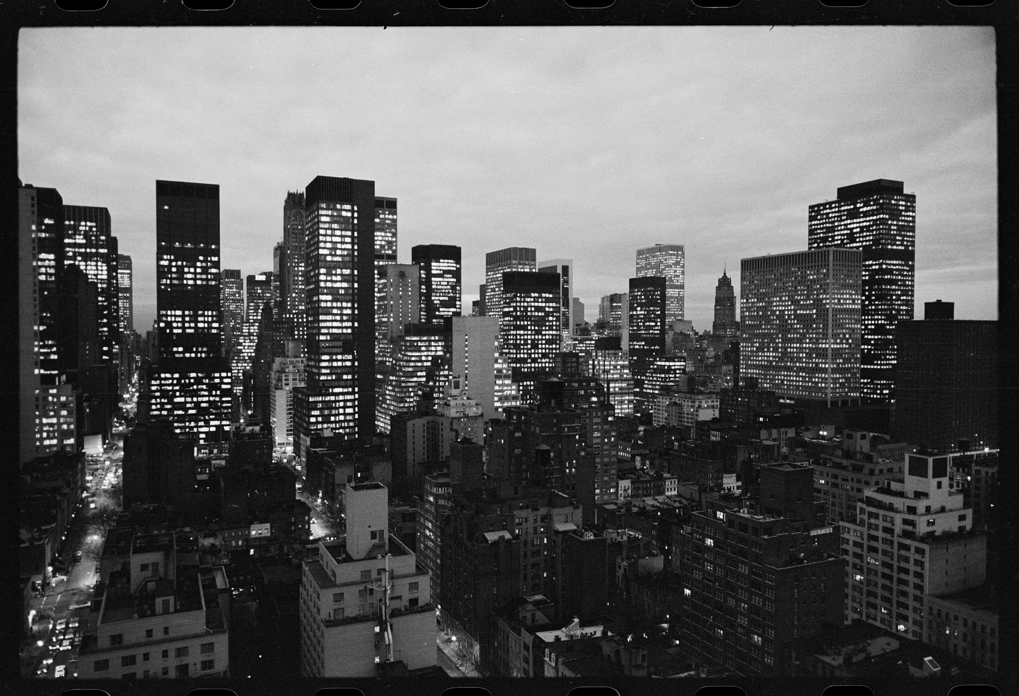 Manhattan buildings seen from above at dusk, in black and white, with office windows lighting up and the streets already dark